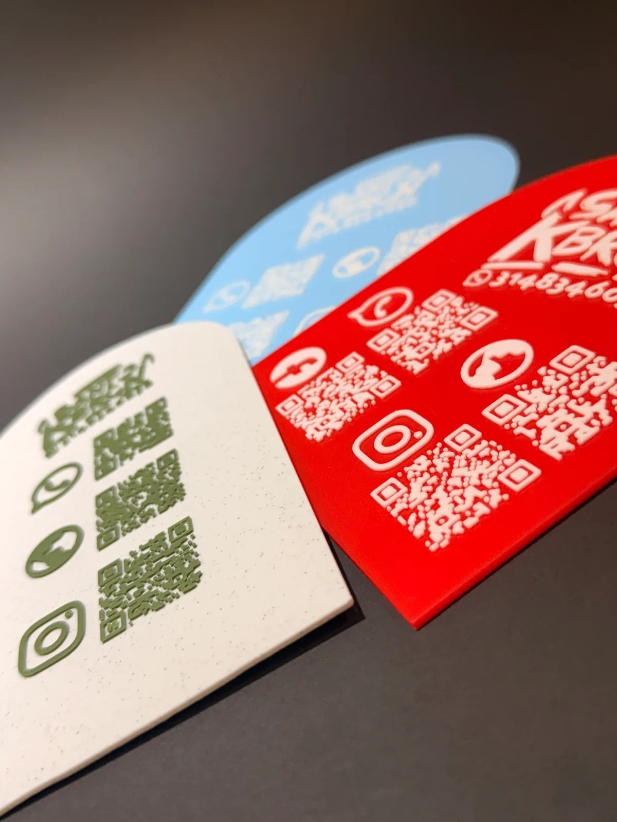 Detalle en primer plano de tres Placas Portátiles QR en crema, azul claro y roja, con los códigos QR y los íconos de redes levantados en relieve sobre la superficie — Placa Portátil QR de Solid Lab 3D, Colombia