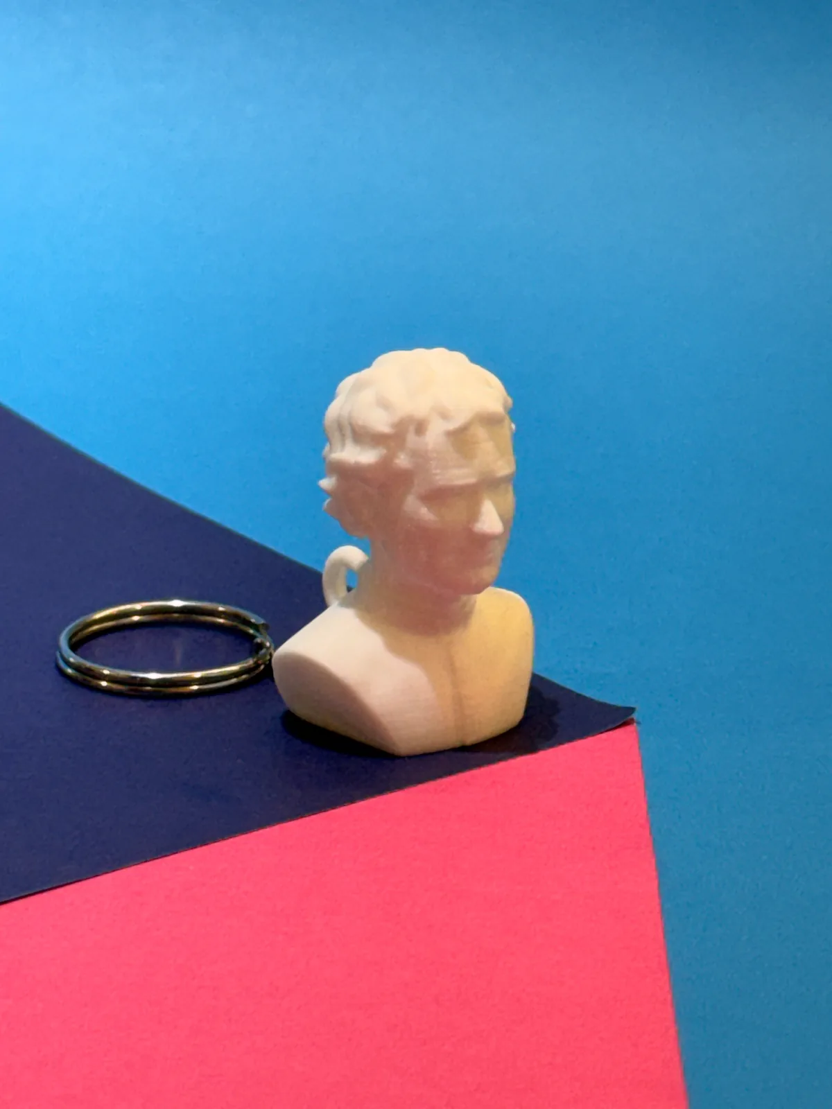 Llavero temático con forma de busto en crema de perfil junto a su argolla metálica, sobre cartulinas azul, azul oscuro y rosada — Llaveros e imanes de Solid Lab 3D, Colombia
