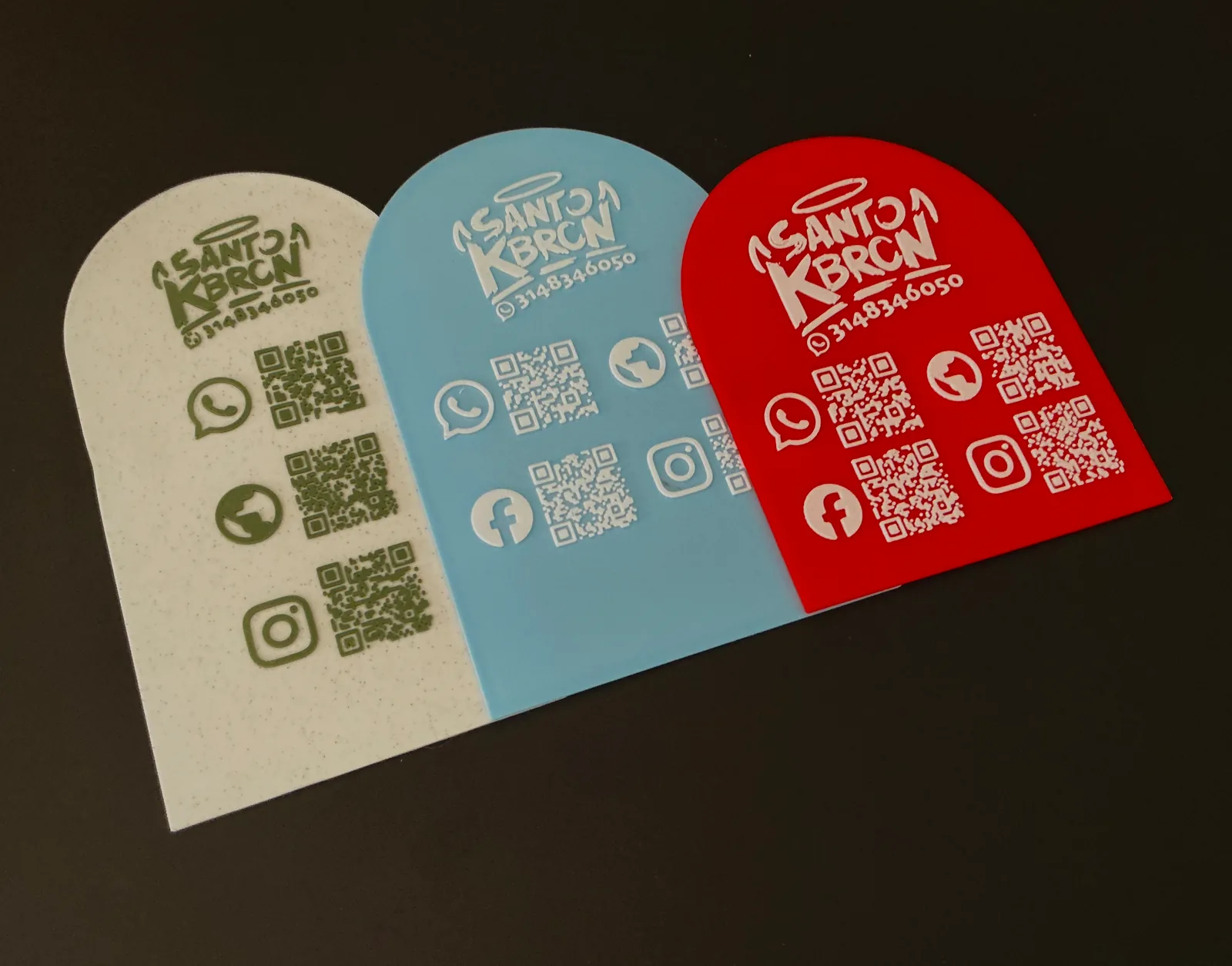 Tres Placas Portátiles QR en crema, azul claro y roja apoyadas sobre una superficie oscura, con el logo del negocio en relieve en la parte de arriba de cada una — Placa Portátil QR de Solid Lab 3D, Colombia